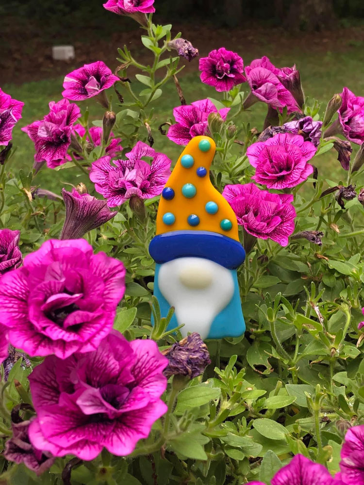 A fused glass gnome plant stake with a colorful orange and blue hat, peeking out from among vibrant pink flowers in a garden.