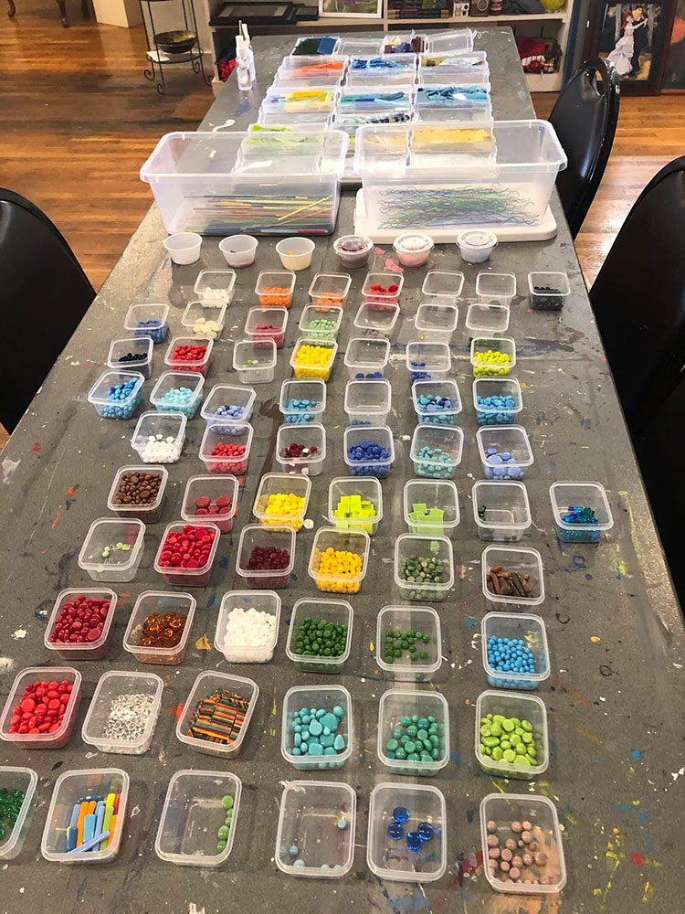 Small plastic containers with colorful glass beads laid out in a grid on a gray table splattered with colorful paint, where art students have clearly been creating.