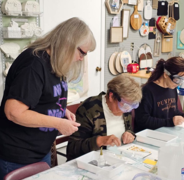 Joanne Fransen-Gilliam, wearing a black shirt with purple text, guides workshop participants working on fused glass projects.