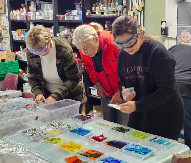 Three workshop participants wearing safety glasses choose colorful glass pieces from trays for fused glass projects.